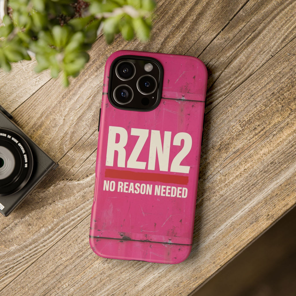 A pink phone case with a distressed, weathered texture and the text 'RZN2 NO REASON NEEDED' in white, shown on a wooden tabletop next to a camera and green leaves.
